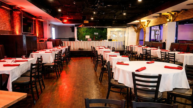 Dimly lit restaurant dining room with white-clothed tables, red napkins, and a stage with a green wall.