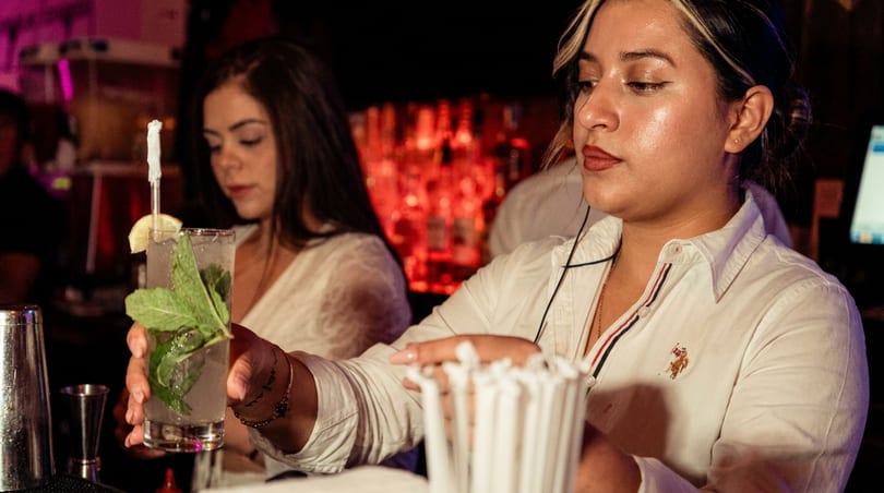 Bartender hands a mojito cocktail garnished with mint and lemon at a lively bar.