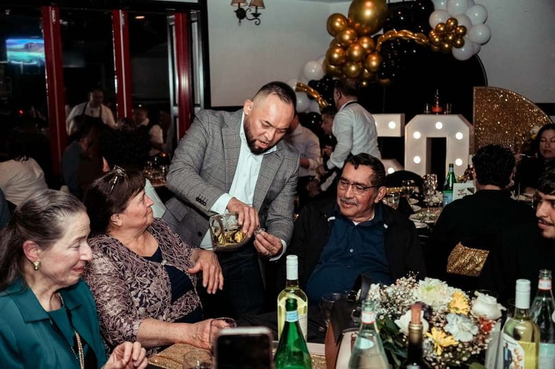 Man in a suit pours a drink for an older man at a lively restaurant celebration.